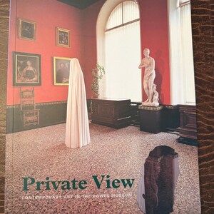 May include: A book titled "Private View" with the subtitle "Contemporary Art in the Bowers Museum" is shown. The cover depicts a room with red walls, artwork, and a draped figure. The book is on a brown surface.