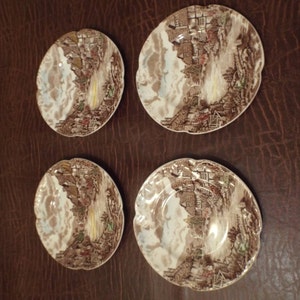 May include: Four vintage brown and white ceramic plates with a landscape design. The plates have a scalloped edge and feature a detailed illustration of a village scene with a waterfall and buildings.