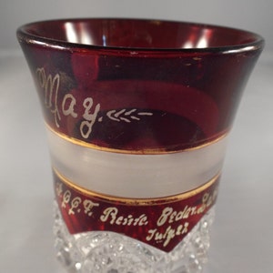 May include: A ruby red glass with a frosted white band and gold trim. The glass is decorated with gold lettering that reads "May" and "1907 F. Reisig, Redern, O. Julper".