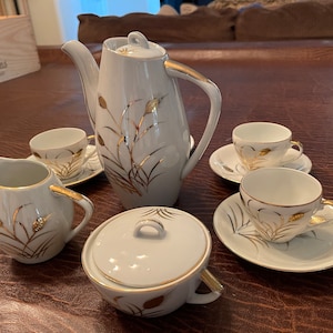 May include: A white tea set with gold accents. The set includes a teapot, creamer, sugar bowl, and four cups and saucers. The tea set features a delicate floral design with gold accents.