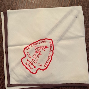 May include: White cotton neckerchief with a red embroidered patch that says "4B Conclave Indian Heritage 1975 Pine Hill S.R. WWW" and depicts two Native American figures dancing.