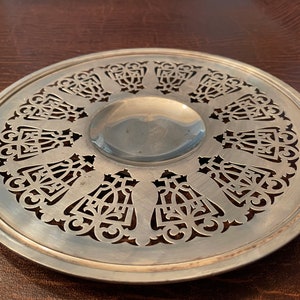 May include: A silver-colored, round metal trivet with an intricate, openwork design. The trivet features a central, slightly recessed area and a decorative border with repeating, stylized floral or geometric patterns. The surface has a brushed finish.