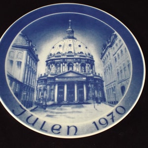 May include: A blue and white decorative plate featuring a detailed illustration of a domed building with columns, likely a cathedral, set within a city scene. The plate is inscribed with the word "Julen" and the year "1970".