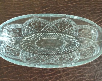 May include: A clear glass oval serving dish with a decorative, cut-glass pattern. The pattern features a central circle with radiating lines and a border of floral motifs.