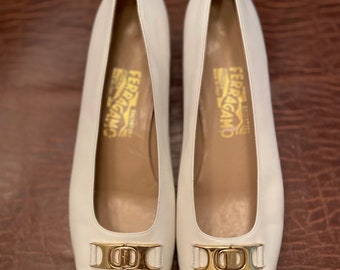May include: A pair of white leather heeled shoes with a beige toe and a gold buckle. The shoes have the text "FERRAGAMO" written on the inside.
