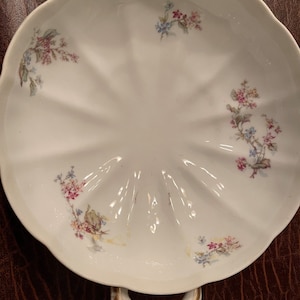 Może przedstawiać: Biała porcelanowa miska do serwowania z dwoma uchwytami i wzorem kwiatowym. Miska ma falisty brzeg i jest ozdobiona delikatnymi różowymi, niebieskimi i zielonymi kwiatami.