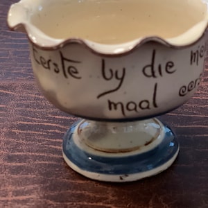 Pode incluir: Um ovo de cerâmica branca com uma borda azul e marrom. A xícara tem uma borda festoneada e as palavras "Eerste by die maal" escritas no lado.