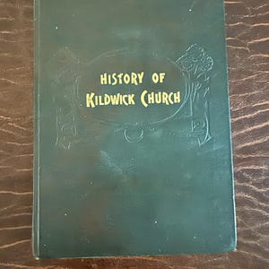May include: A dark green book with the title "History of Kildwick Church" in gold lettering. The book has an embossed design and is on a brown, textured surface.