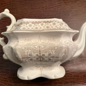 Op de afbeelding: Witte keramische theepot met een decoratief bloemen- en geometrisch patroon in grijstinten. De theepot heeft een gebogen handvat, een tuit met een vogelachtig ontwerp en een deksel met een bijpassend patroon.