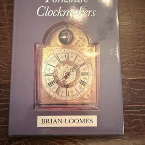 Peut inclure: Livre intitulé "Yorkshire Clockmakers" avec une illustration de cadran d'horloge sur la couverture. La couverture est de couleur violet clair, avec le nom de l'auteur, Brian Loomes, imprimé en bas.