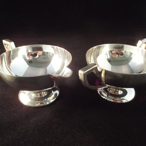 May include: A set of two silver creamer and sugar bowls with a rounded shape and a single handle. The bowls have a polished finish and a simple design.