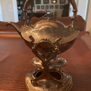 May include: A silver teapot with an ornate design and a stand. The teapot has a lid with a knob on top and a handle that curves up and over the top of the teapot. The stand has three legs and a round base.