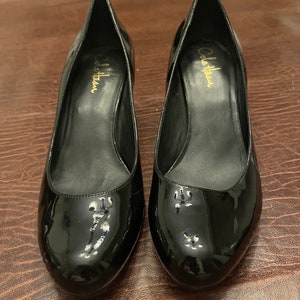 May include: A pair of black patent leather Cole Haan pumps. The shoes have a pointed toe and a mid-height heel. The Cole Haan logo is visible in gold lettering on the insole. The shoes are displayed on a brown surface.