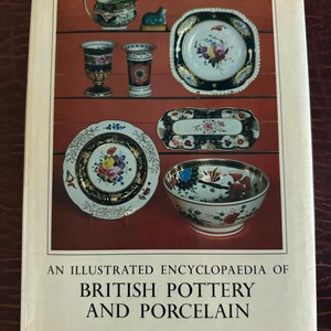 May include: An Illustrated Encyclopaedia of British Pottery and Porcelain book cover. The cover features a collection of antique pottery and porcelain pieces, including plates, bowls, and vases, with floral designs. The book is a second edition by Geoffrey A. Godden.