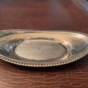 May include: A silver-colored, oval-shaped serving tray with a decorative rope-like border. The tray's interior features an embossed floral design. The tray is set against a brown surface.