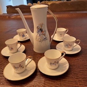 May include: A white porcelain tea set with gold trim. The teapot features a colorful illustration of a couple in a cafe. The cups and saucers have matching illustrations of people dancing.
