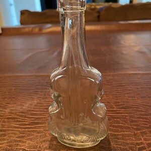 May include: A clear glass bottle shaped like a violin. The bottle is empty and has a smooth, rounded surface.
