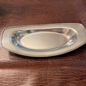 May include: A rectangular, silver-colored serving tray with an oval interior. The tray has a decorative border and small emblem on each end. The tray is displayed on a brown, textured surface.