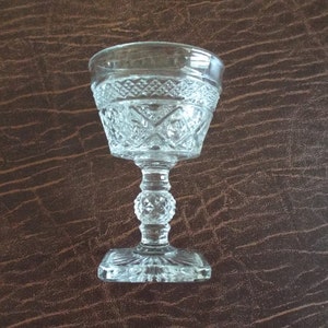 May include: A clear, vintage-style glass goblet with a decorative diamond pattern on the bowl. The goblet has a round, beaded stem and a square base. The glass is set against a brown background.