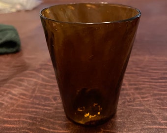 May include: A brown glass tumbler with a slightly flared rim. The glass has a textured surface and appears to be hand-blown.