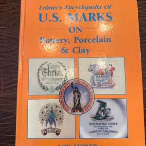 Puede incluir: Un libro naranja titulado "Lehner's Encyclopedia Of U.S. Marks On Pottery, Porcelain & Clay". La portada muestra marcas de cerámica y el nombre de la autora, Lois Lehner. Es una guía de referencia para coleccionistas.