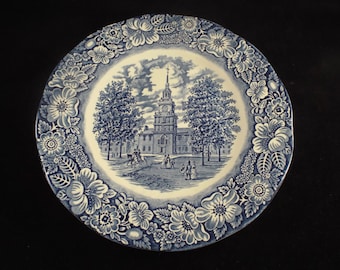 May include: Blue and white ceramic plate with a floral border and a central image of a building with a steeple. The building is surrounded by trees and people.