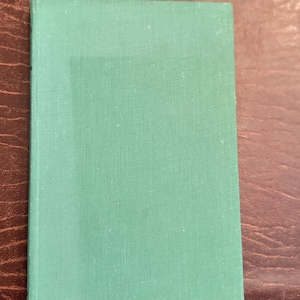 Könnte beinhalten: Ein rechteckiges Hardcover-Buch mit einem einfarbigen grünen Einband. Der Einband hat ein leicht strukturiertes Aussehen und einen helleren grünen rechteckigen Bereich in der Mitte. Das Buch liegt auf einer braunen Holzoberfläche.
