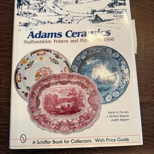 Op de afbeelding: Een boek getiteld "Adams Ceramics" met afbeeldingen van decoratieve borden. De omslag toont een landschapsillustratie en de tekst "Staffordshire Potters and Pots, 1779-1998". Het boek is een Schiffer Book for Collectors.