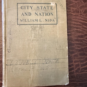 May include: A vintage book titled "City State and Nation" by William L. Nida. The cover is a faded tan color with black text in a rectangular box. Handwritten inscriptions are visible on the cover.