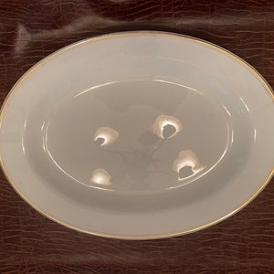 Puede incluir: Plato de servir ovalado gris claro con borde dorado. El plato tiene un centro ligeramente elevado con cuatro pequeñas hendiduras redondeadas. El diseño es simple y elegante, adecuado para servir alimentos.