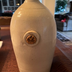 以下が含まれることがあります： 丸みを帯びたボディと細い首を持つベージュ色の炻器のジャグ。コルクストッパーと円形のエンブレムが特徴です。このジャグは素朴で手作りの外観をしており、ヴィンテージまたはアンティーク品であることを示唆しています。