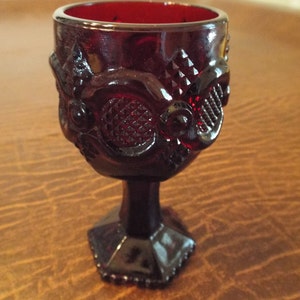 May include: A ruby red glass goblet with ornate, raised floral and geometric patterns. The small, stemmed glass has a hexagonal base and a detailed design around the bowl. The glass is approximately 3 inches tall.