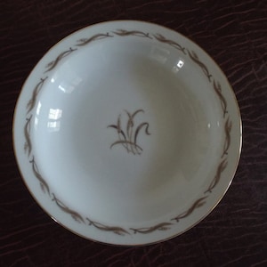 May include: A white ceramic bowl with a gold rim and a wheat stalk design in the center. The rim features a repeating brown wheat stalk pattern. The bowl is likely for serving food.