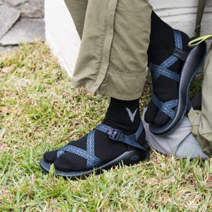 May include: Black sandals with blue patterned straps are worn with black socks. The sandals have a toe separator and adjustable straps. The person is wearing olive green pants and is sitting on the grass.
