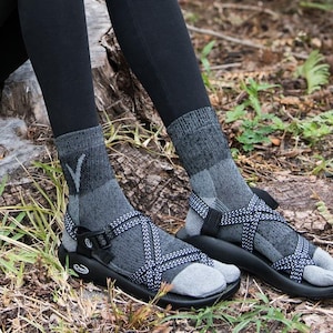 May include: Close-up of a person's feet wearing gray socks and black sandals with crisscross straps. The sandals have a toe separator and are on a black sole. The person is sitting on a tree stump surrounded by foliage.