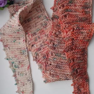 May include: A hand-knitted scarf in shades of cream, orange, and red with a speckled pattern. The scarf has a textured, open weave design and a scalloped edge. The scarf is made of soft yarn and is suitable for cooler weather.