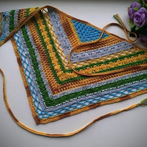 May include: A handmade crochet shawl in a triangular shape. The shawl features a vibrant mix of colors, including blue, green, yellow, brown, and gray. It has a textured design with a border and long ties with tassels.