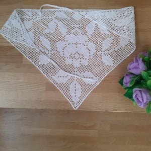May include: A white, triangular crochet shawl with a floral pattern. The shawl is laid out on a wooden surface. The crochet work is detailed, and the shawl has a tie at the top. A bouquet of purple flowers is in the background.