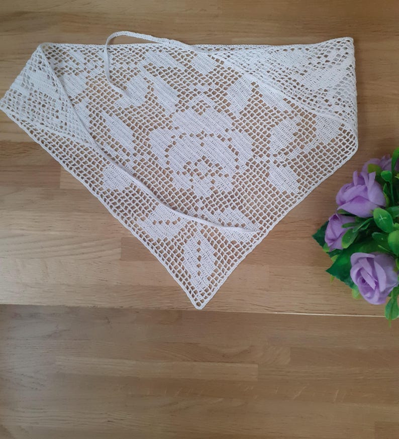 May include: A white, triangular crochet shawl with a floral pattern. The shawl is laid out on a wooden surface. The crochet work is detailed, and the shawl has a tie at the top. A bouquet of purple flowers is in the background.