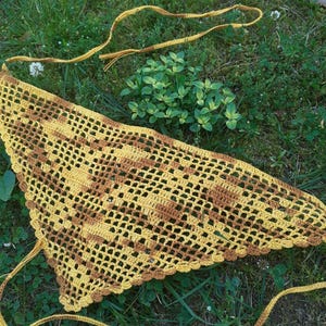 May include: A handmade, yellow and brown crochet shawl with a triangular shape. The shawl features a detailed openwork design and a scalloped edge. Long, yellow ties extend from the corners. The shawl is displayed on a bed of green grass.
