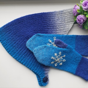 May include: A knitted blue and grey scarf and matching mittens set. The scarf has a button fastening and a gradient colour effect. The mittens are blue with white snowflake embroidery. The set is made of a soft, warm material.