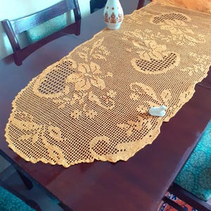 May include: An oval-shaped, golden-yellow crochet table runner featuring an intricate openwork pattern of floral motifs and geometric grid sections. The runner has a scalloped edge and is laid flat on a dark brown wooden surface.