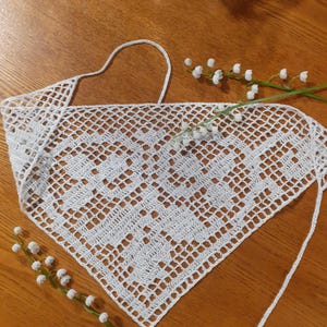 May include: A white triangular crochet bandana featuring an intricate openwork grid pattern with solid block motifs. The accessory includes thin crocheted ties at the top corners and is displayed alongside two sprigs of white lily of the valley flowers.