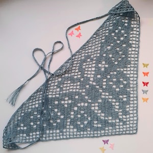 May include: A triangular crochet bandana in a dusty blue hue, featuring an intricate openwork grid pattern and delicate floral motifs. The accessory includes thin, matching blue ties with small tassels at the ends for securing the garment.