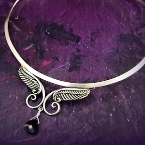 Puede incluir: Un collar choker plateado con un delicado diseño de alas y una piedra preciosa negra colgante. El collar está sobre un fondo morado.