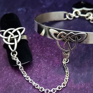 May include: A silver Celtic knot ring and bracelet connected by a chain. The ring and bracelet both feature a triangular knot design.