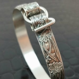 Puede incluir: Un brazalete de plata con un diseño floral y un cierre de hebilla.