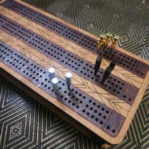 May include: A rectangular wooden cribbage board featuring alternating light and dark wood grain sections with laser-engraved geometric patterns. The board includes three rows of holes for scoring and nine metal pegs in silver, black, and gold.