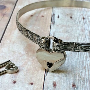 Puede incluir: Un collar choker plateado con un candado en forma de corazón y una llave a juego. El collar está decorado con diseños florales.