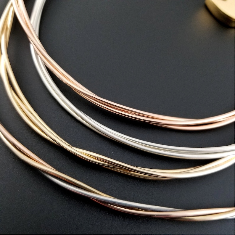 May include: A set of four intertwined metal rings in gold, silver, and rose gold tones. The rings are arranged in a circular pattern, creating a layered and textured effect.